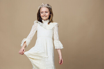 Young girl wearing a white lace dress and a crown, smiling joyfully against a neutral background,...