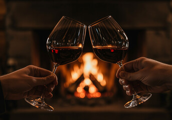 Wine glasses with red wine cheers in front of fireplace with warm light. Two wine glasses toasting with red wine in cozy atmosphere