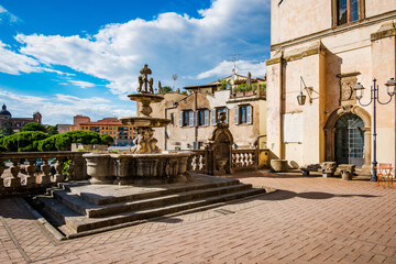Ancient fountain standing in Piazza della Rocca, Viterbo, Italy, casting shadows on the brick pavement, with historic buildings and a glimpse of the cityscape under a bright sky