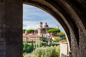 The beautiful cityscape of Viterbo, Italy, with its church and traditional houses, is framed by an ancient stone arch, creating a picturesque view of the city in the Lazio region