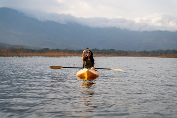 Latin hispanic female navigating on a serene lake with a kayak, having fun taking a selfie as...