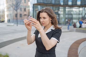 A young woman in a black dress with a white collar looks in a small mirror. She has long brown hair...