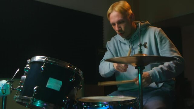 Young blond drummer in blue hoodie passionately hits drums, drummer focused on rhythm, musician mastering drums, drummer practicing beats, energetic musician playing drums in studio.