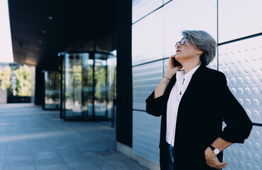 Confident senior Caucasian businesswoman with gray hair making important phone call near modern...
