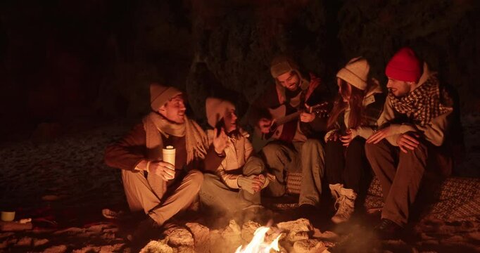 Guy in winter clothes and a hat with earflaps plays the guitar while his friends laugh and chat while sitting near the fire on a cozy evening on the beach