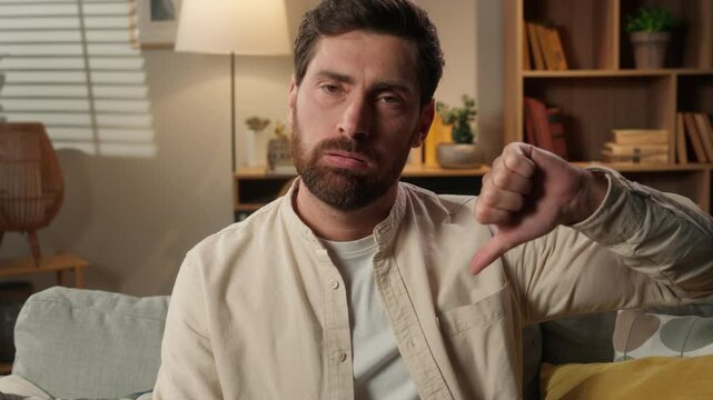 Sad frowning upset man Caucasian dissatisfied guy male home owner on couch showing thumb down gesture finger dislike hand gesture bad sign disapprove no negative result displeased looking at camera
