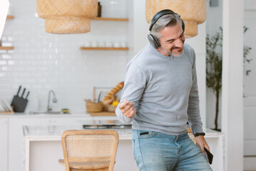 Happy Middle age Caucasian man listen to music in headphones use mobile cell phone in living room at home, choosing favorite energetic disco music in mobile application. Happy enjoy life concept.