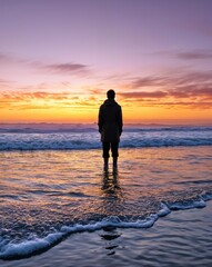 A solitary figure stands partially submerged in calm waves at the shoreline, gazing at a vibrant sunset that paints the sky with hues of orange and purple, creating a serene atmosphere of tranquility.