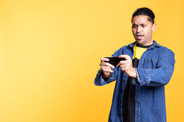 Cheerful indian person playing video games on his mobile phone, celebrating after defeating foes against orange background. Young player with man bun, thrilled about online games.