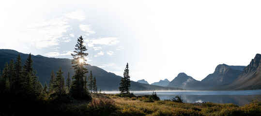 Panorama of calm lake, mountains and trees looking at the sun with a partially transparent sky