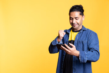 Happy gamer celebrating after winning game on gaming console, standing against studio background. Delighted Indian man bragging after being victorious in video gaming competition, defeating enemies.