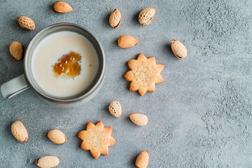 A warm cup of creamy coffee sits beside flower-shaped almond cookies, surrounded by whole almonds on a textured gray surface, creating a delightful snack arrangement