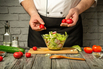 A chef in a black apron prepares a fresh salad by adding radishes to a bowl of mixed greens on a rustic wooden table in a well-lit kitchen