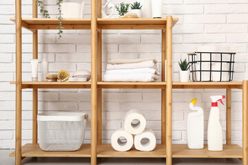 Wooden shelving unit with toilet paper rolls, bath supplies and detergents near light brick wall