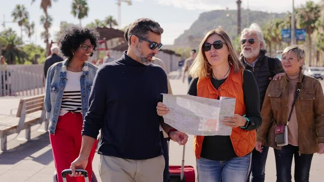 Group diverse tourist people walking with suitcases together looking paper map enjoying sunny autumn vacations. Adults friends strolling talking happy sightseeing in city center on holiday travel