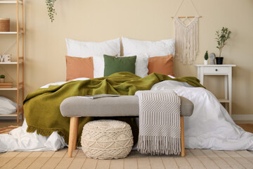 Big comfortable bed with crumpled linen, shelving unit, bedside table and bench in interior of bedroom