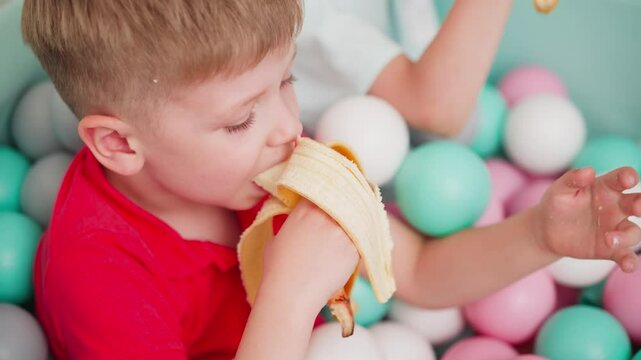 Aerial view of little kids sitting in pastel ball pit enjoying bananas together, boy in red shirt focused on snack while colorful balls surround them, joyful and playful indoor moment