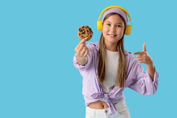 Cute teenage girl in headphones pointing at delicious cookie on blue background