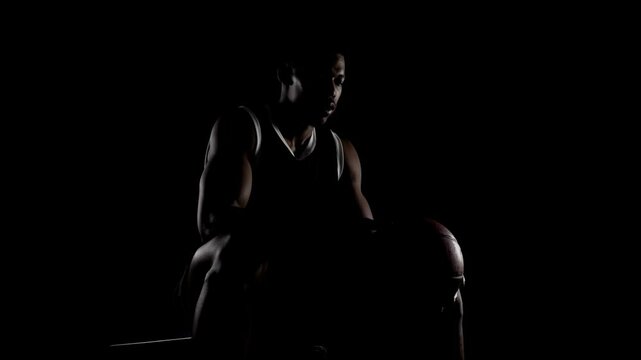 Basketball player sitting on a bench. Concentrated african american man taking deep breath and looking down against black background.