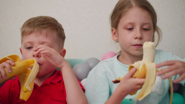 Brother and sister sitting in pastel ball pit peeling and eating bananas, boy wearing red shirt and girl in cyan top focused on fruit, natural candid moment with playful atmosphere