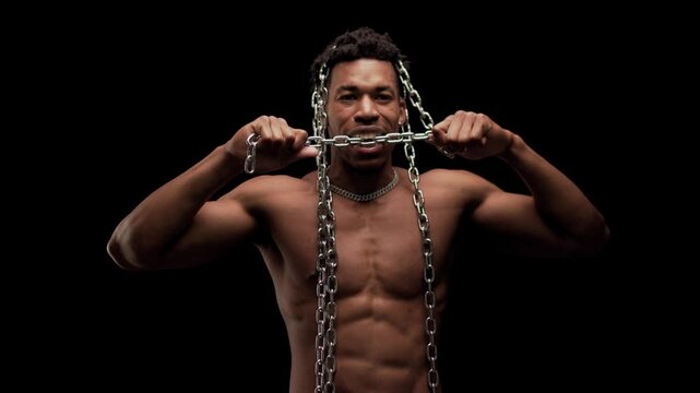 Muscular man showing muscles and biting a metal chain. African American male posing against black background