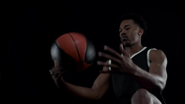 Basketball player balancing a ball. African american sports man sitting and practicing against black background.