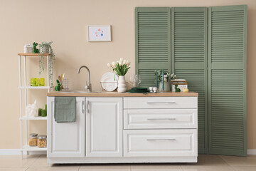 Interior of festive kitchen with folding screen, utensils, flowers and Easter decorations on counter near beige wall