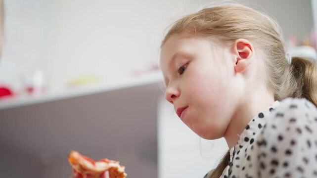 Young kid with hair tied back wearing spotted clothes eagerly bites into pizza slice indoors with soft natural light, partial view of another person nearby