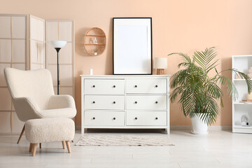 White chest of drawers with blank photo frame and comfortable armchair in room