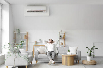 Fototapeta premium Young man resting on sofa with air conditioner in living room
