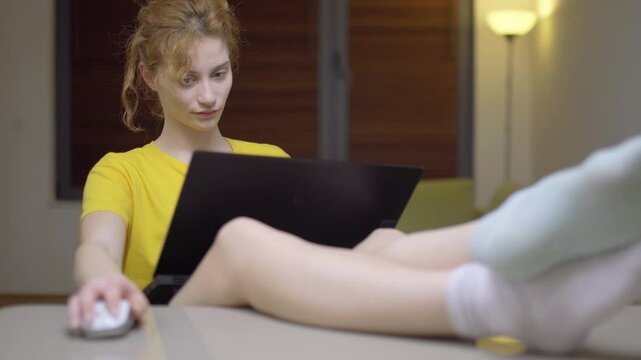 Girl working on a laptop in her lap. Casual clothing different color socks. Feet up on the table in a living room at home. Portrait of a student taking online course. Home education. Left camera
