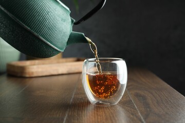 Pouring freshly brewed tea from teapot into cup at wooden table, closeup
