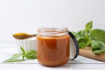Tasty curry sauce in glass jar, powder and basil on white wooden table, closeup