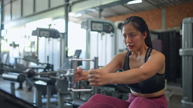 Focused asian sportswoman performing seated cable rows, pulling cable handles toward chest, targeting back muscles with intense concentration inside modern fitness facility