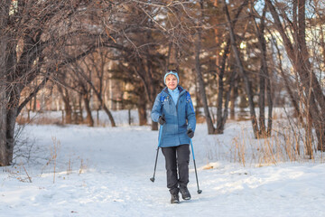 Mature woman in winter park with Nordic walking poles, enjoying the tranquility of nature and improving her health. Active lifestyle concept, winter walks, outdoor fitness.