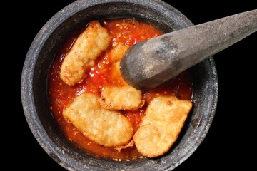 Ingredients for making Chili Sambal, hot red chili sauce and fried tempe on a mortar made of stone and pestle isolated on black background.

