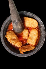 Ingredients for making Chili Sambal, hot red chili sauce and fried tempe on a mortar made of stone and pestle isolated on black background.
