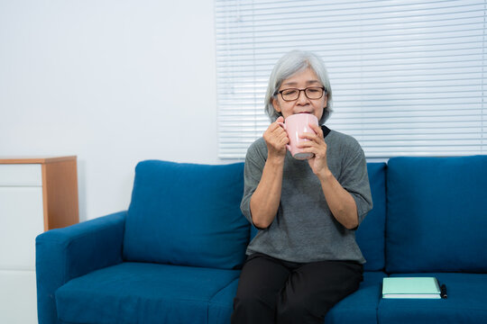 Elderly Asian woman with grey hair and eyeglasses sipping a hot beverage from a pink mug while relaxing on a blue sofa in her cozy living room, with a notebook and pen nearby - Powered by Adobe