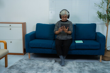 Elderly asian woman wearing headphones is using a tablet while sitting on a comfortable blue sofa in her modern living room, enjoying technology and staying connected in retirement