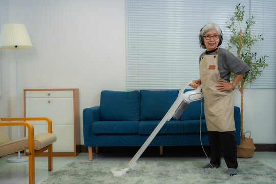 Elderly asian woman listening music with headphones and vacuuming carpet in living room, she's wearing an apron and is posing with hand on hip