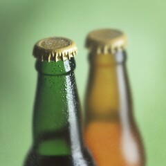 Close up of two beer bottles with condensation, one green and one amber, against a blurred green background. The bottle caps are gold colored