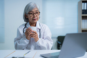 Senior female doctor wearing lab coat and stethoscope is holding a coffee cup while sitting at desk...