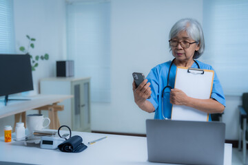 Senior female doctor wearing blue uniform and stethoscope is using smartphone and holding clipboard while standing in hospital office with medical equipment on desk