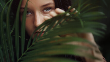 A close-up shot of a woman face as she peers through lush tropical foliage, her eyes partially...