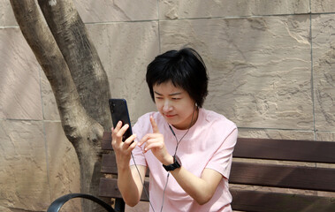 A short-haired Asian woman wearing a pink cotton t-shirt and sitting on a park bench making video calls on her smartphone.
