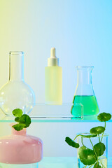 Dropper serum bottles placed on a glass podium, behind are Florence flask and conical flask containing green liquid. The essence extracted from natural pennywort is green. Vase with centella plants.