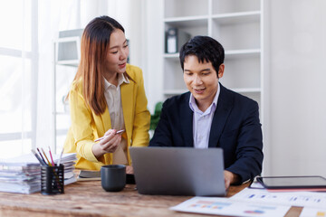 Two happy asian employees working together using computer planning project. business woman consulting teaching young employee looking at laptop sitting at desk in office
