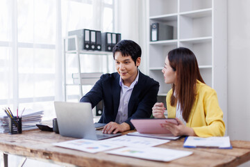 Two happy asian employees working together using computer planning project. business woman consulting teaching young employee looking at laptop sitting at desk in office
