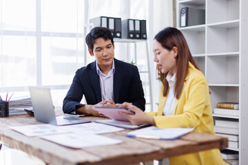 Two happy asian employees working together using computer planning project. business woman consulting teaching young employee looking at laptop sitting at desk in office
