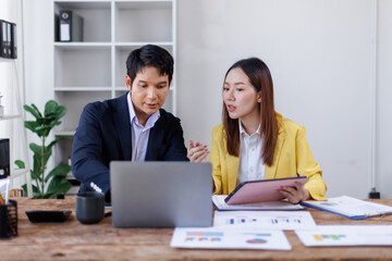 Two happy asian employees working together using computer planning project. business woman consulting teaching young employee looking at laptop sitting at desk in office
